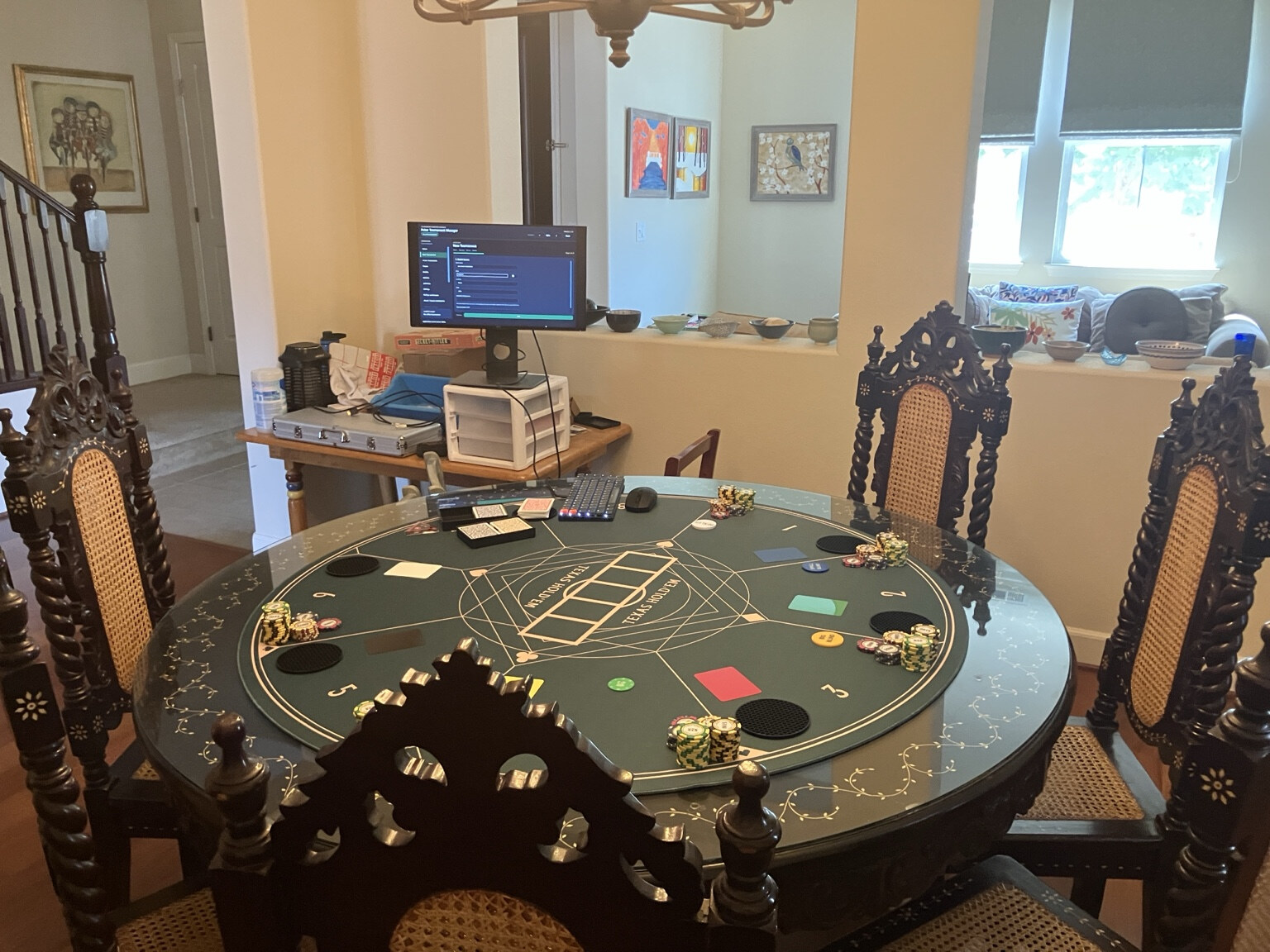 Physical poker tournament setup with a round table, chips, laptop controls, and an external application display positioned for the host.